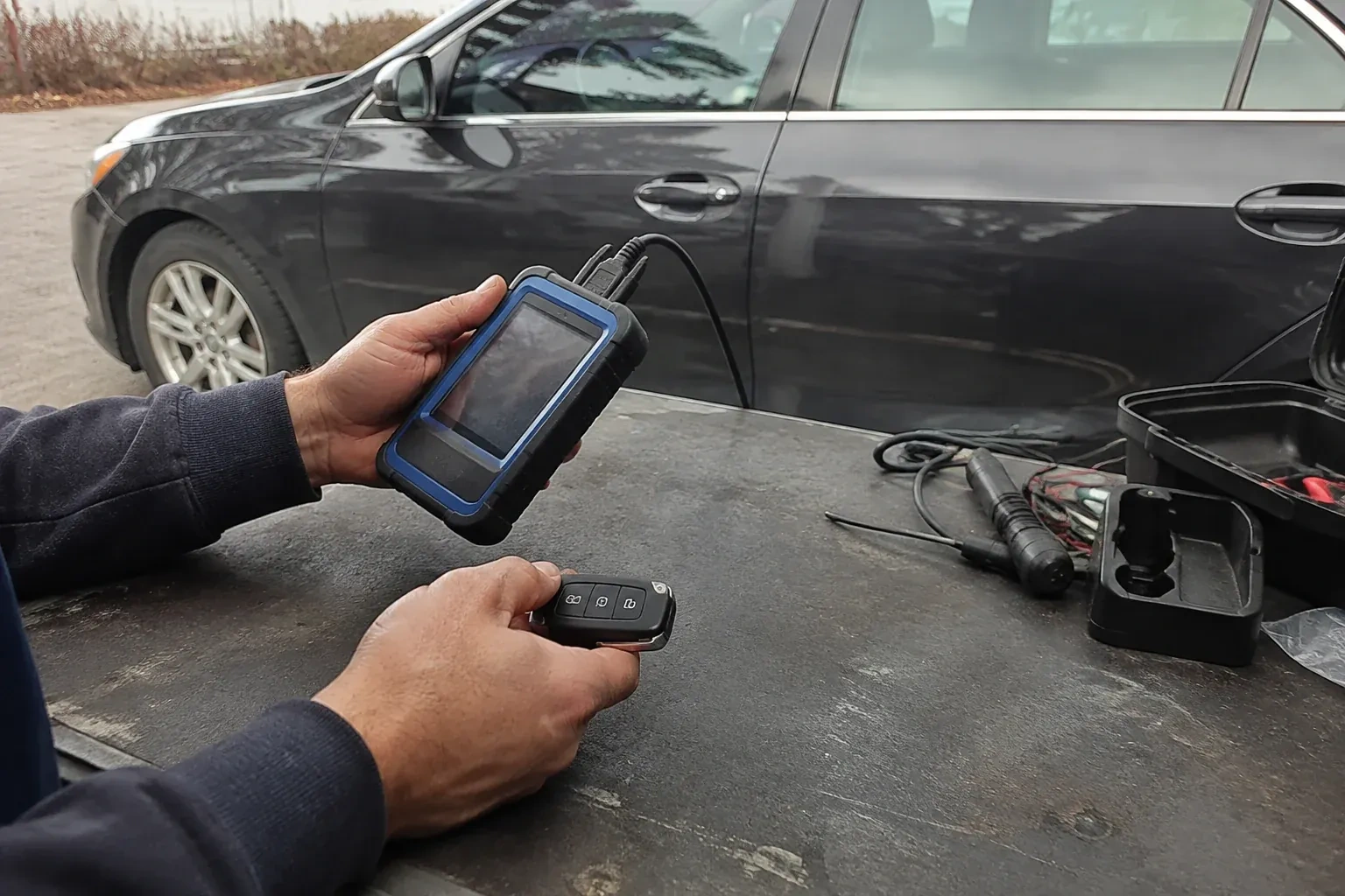 Hands holding a blue-edged electronic device and car-key fob at a worktable beside a parked car. — Transponder Key Programming, Wilmot