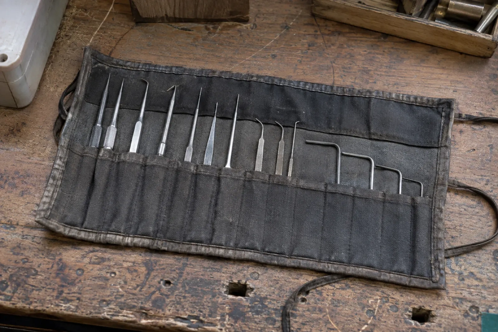 Roll-up black cloth pouch lying open on a worn wooden bench, holding thin metal probes and hooked tools in separate pockets.